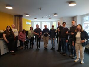 A photo of the Working Group members: This photo captures a group of people standing together in a room. The individuals are standing side by side, smiling, and seem relaxed. Some members of the group are holding tactile writing devices, possibly Braille devices or tools, while a couple of people are using white canes. One person is accompanied by a guide dog.
The room has yellow walls and large windows in the background that allow natural light to fill the space. The decor is modern. There are shelves with possibly motivational phrases.
The mood of the image is friendly and inclusive, with the group appearing to enjoy each other’s company. The setting seems like an accessible workspace or meeting venue, likely accommodating the needs of visually impaired participants.