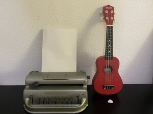 a perkins machine with the paper rolled in, beside it an ukulele with braille sticker and a pick