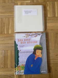 The German braille release and the standard version of the Spanish book The village of stones (Das Dorf der Steine/Hablan las Piedras) by Lawrence Schimel and Lena Studer. The books are both closed and are placed on a wooden floor. The cover of the standard release of the book in the bottom half of the picture shows a drawing of a girl hugging a gravestone. She is smiling and has her eyes closed. She wears a thick blue coat, a red scarf, and a green hat. In the background there are autumn leaves, a squirrel and a bird, some flowers, and a bush in front of a fence. The cover of the braille version in the top half made of a translucent plastic sheet that reveals a shape made out of braille dots on the first page underneath. The shape is partially covered though by a braille sticker and a sticker with roman letters both reading the title of the book. The braille version is a ring binding book. 

