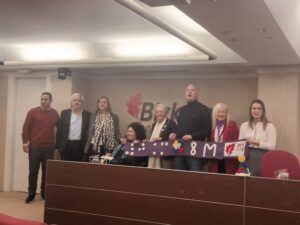 A group foto of 8 people standing behind the desk on the stage in the conference room. Together they are holding the purple banner from the previous picture.