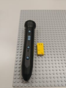 A black penfriend pen and a yellow lego braille brick with the letter e on a grey lego ground plate.