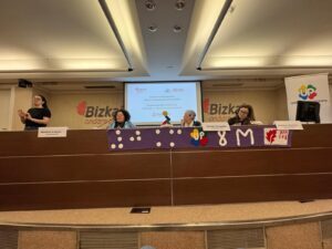 Four women seated at a desk on the stage of a conference room giving a presentation. In front of their desk there is a banner made out of purple wool with crocheted letters and logos. From left to right: “8KM” as white braille, the logo of the “Union Progresista” of ONCE (the letters U and P on four red, blue, yellow, and green hearts forming a cloverleaf), “8M” as white letters, and the logo of the administration of the basque region of Bizkaia (a red leaf on white background). Behind the women there is the projected slide of a presentation with the title “Women with disability: Weaving networks to prevent unwanted loneliness”, written in both spanish and basque.